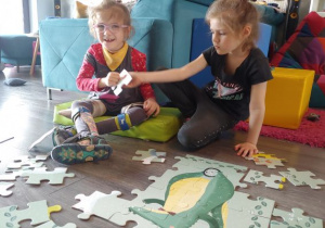 dziewczynki siedzą w domu, na podłodze, obok niebieskiej kanapy i układają duże puzzle